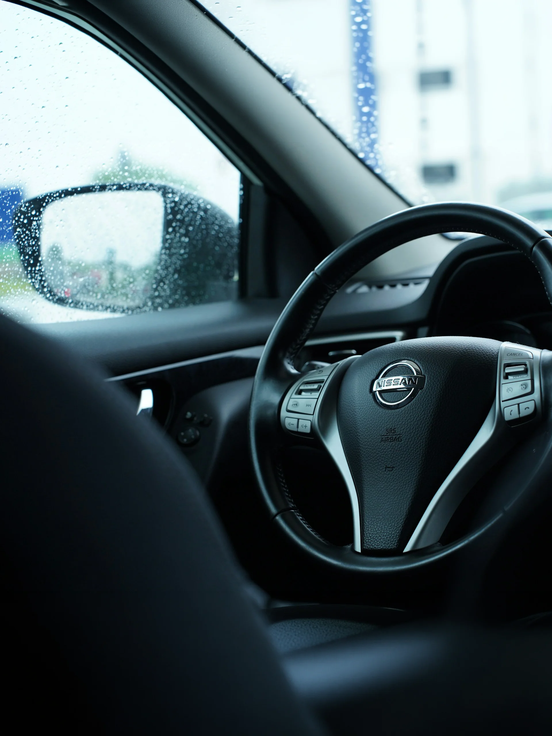 Nissan Certified Collision Repair Interior Systems Nissan steering wheel inside a vehicle with rain-speckled window and side mirror.